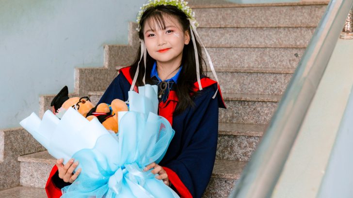 Happy young graduate smiling on stairs with bouquet and stuffed toy.