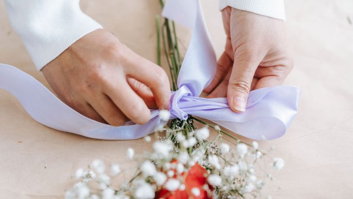From above of crop unrecognizable person tying light ribbon on flowers against light table in daytime