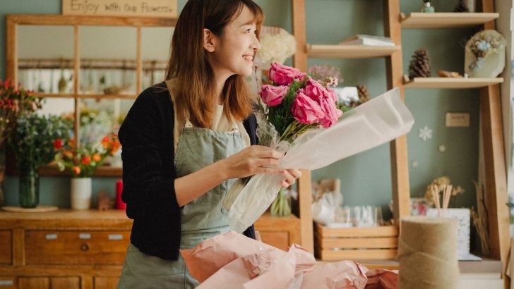 Optimistic Asian female florist wearing apron making flower bouquet with roses while standing in floral workshop