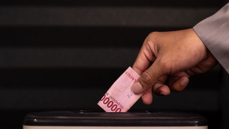 Close-up of a hand placing a 100,000 Indonesian Rupiah note into a donation box, highlighting charity and giving.