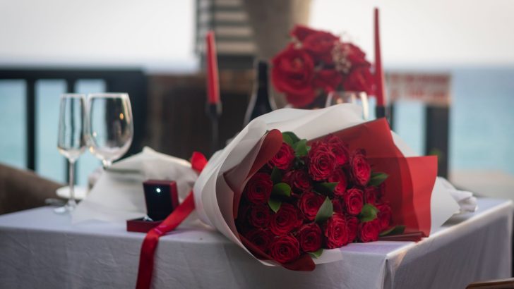 A romantic dinner setup featuring a bouquet of red roses, glasses, and ring box overlooking the sea.
